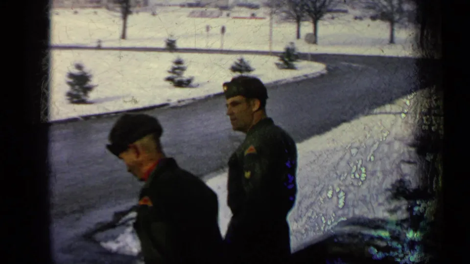 Two soldiers in dark green uniforms walk down exterior steps at a snowy Germany military base, with road and trees behind them.