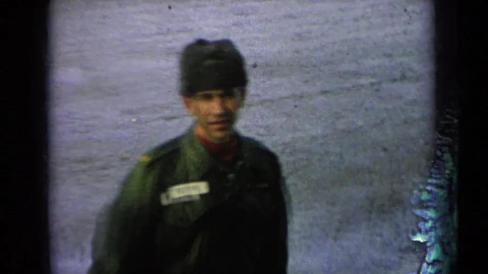 A soldier in a dark green uniform and cap faces the camera, with a name tape visible against the snowy base landscape.