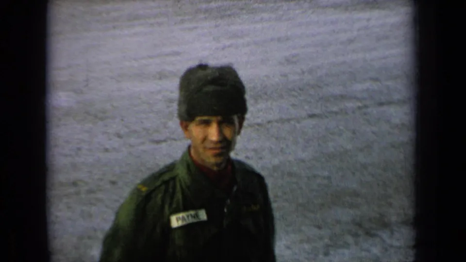A uniformed man wearing a dark winter hat faces the camera, with a name patch and soft 8mm grain visible.