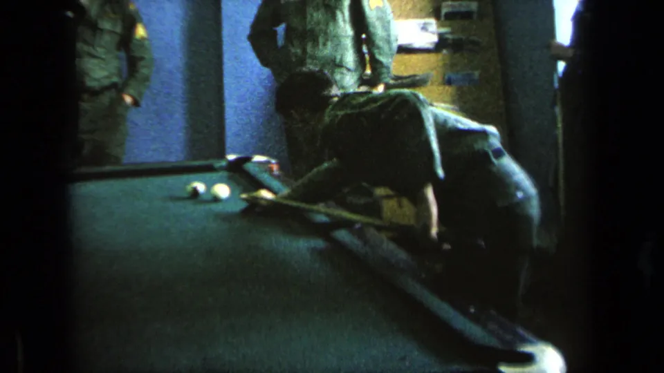 Inside a dim military-base room, a man bends over a green pool table while uniformed men stand nearby.