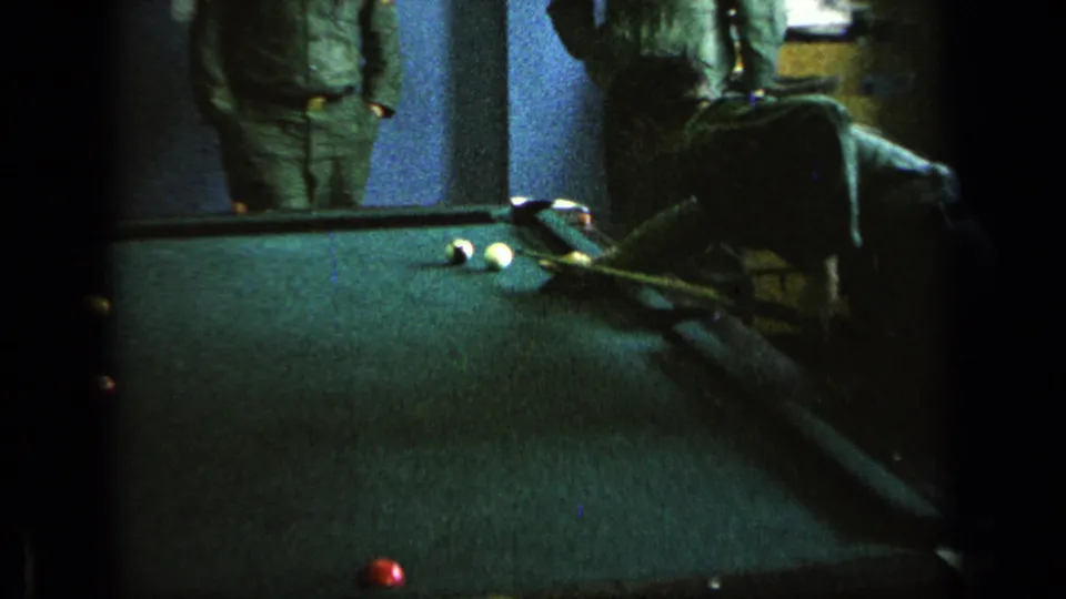A green pool table fills the frame with billiard balls and cues visible while uniformed men stand at the edge of the room.