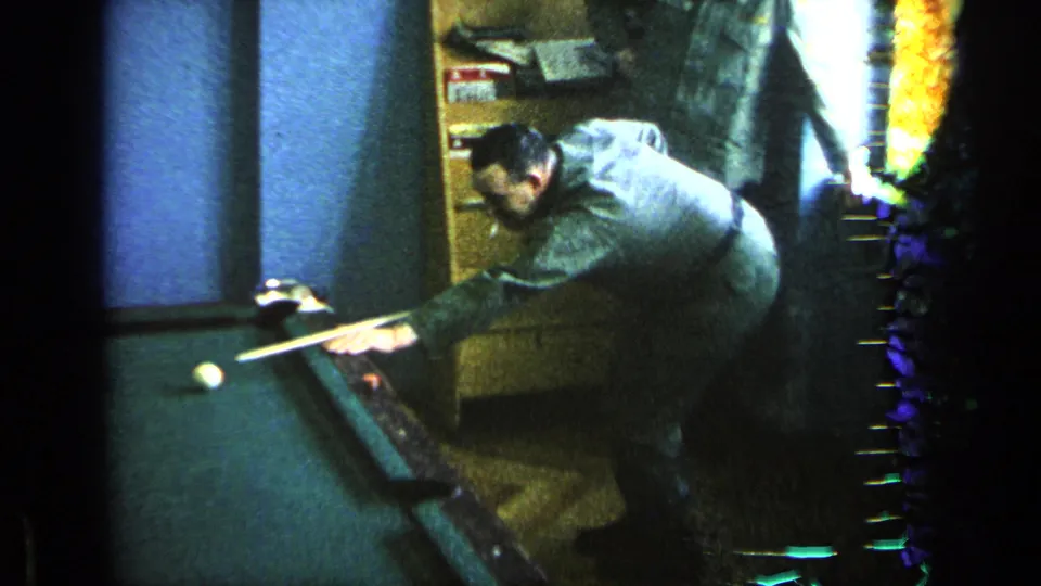 Man in military uniform leans over a green pool table while aiming a cue in a dim indoor room.