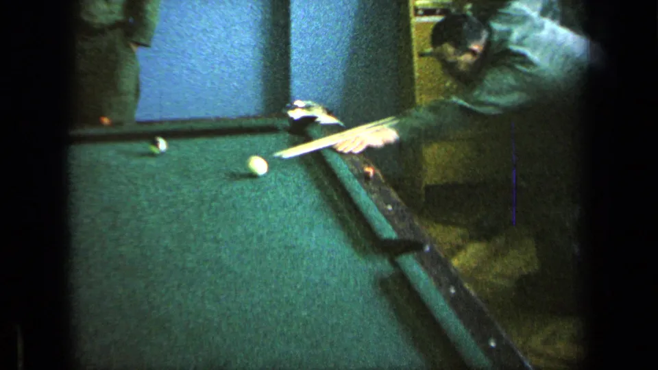 Uniformed pool player aims from the table edge as other service members stand nearby in the recreation room.