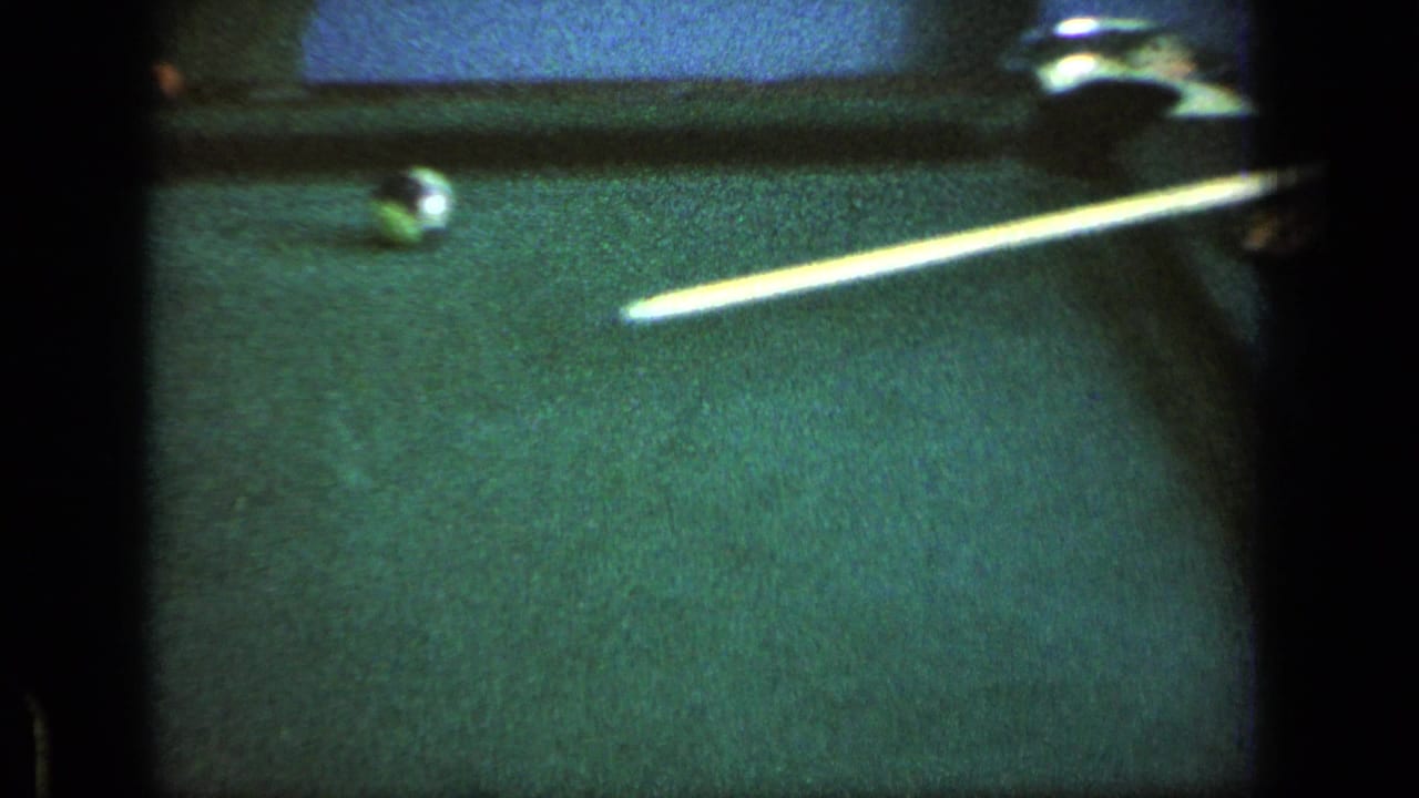 Close view of a green billiards table with a cue stick lined up near the white ball in 1963 8mm base recreation footage.