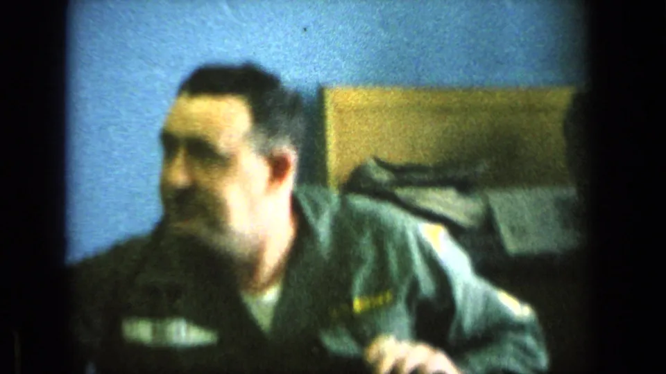 A uniformed adult man appears beside the pool table in an indoor recreation room, with a blue wall and soft background objects visible.
