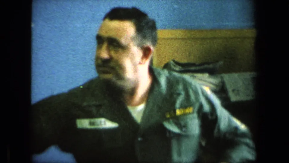 1963 8mm frame of a U.S. soldier in uniform near blue walls and wooden shelving inside a barracks room.