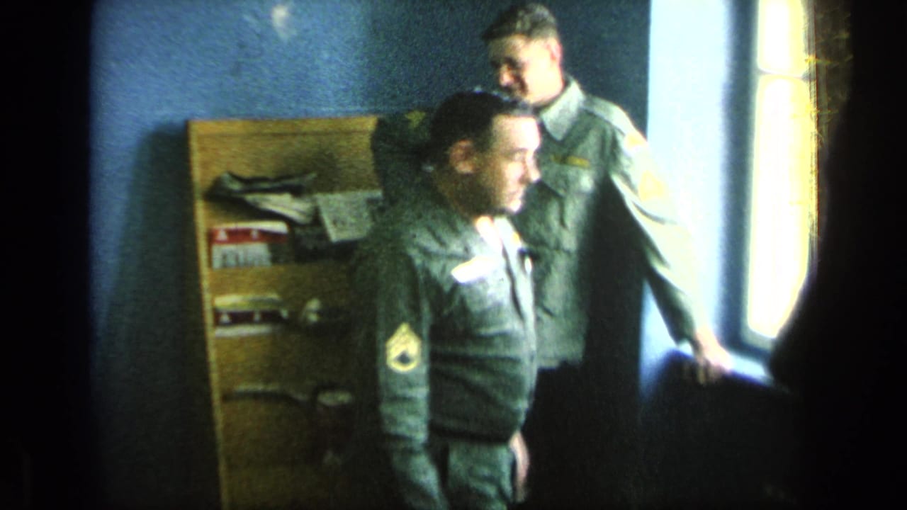 Two uniformed men stand near a bright window in a barracks room.