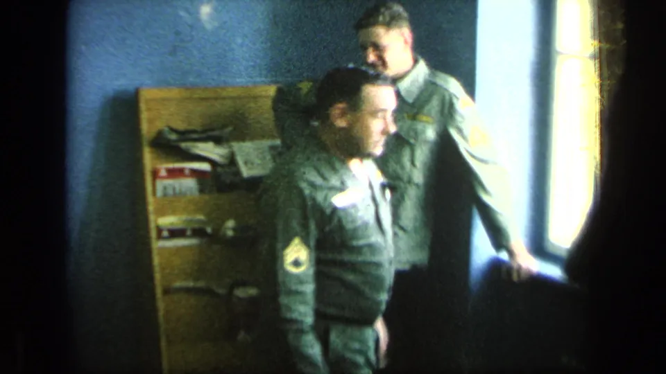 Two U.S. soldiers in uniform stand by a bright window beside wooden shelving in the barracks interior.