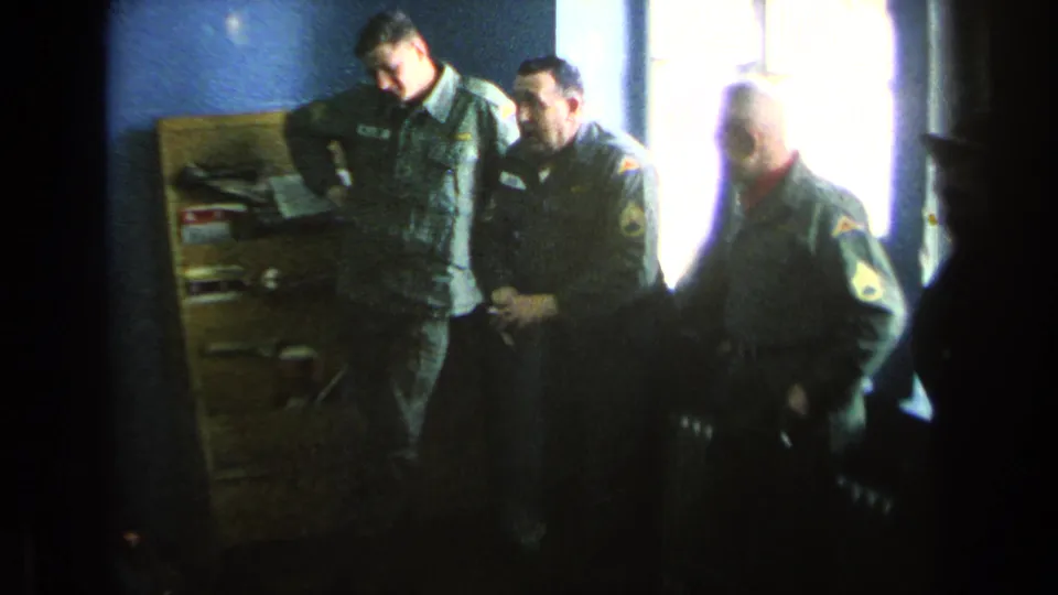 Three U.S. soldiers in uniform stand together inside a barracks room with blue walls and a bright window.