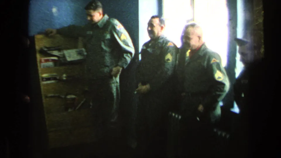 Three uniformed U.S. soldiers appear to stand at attention inside a blue-walled barracks room in 1963 footage.