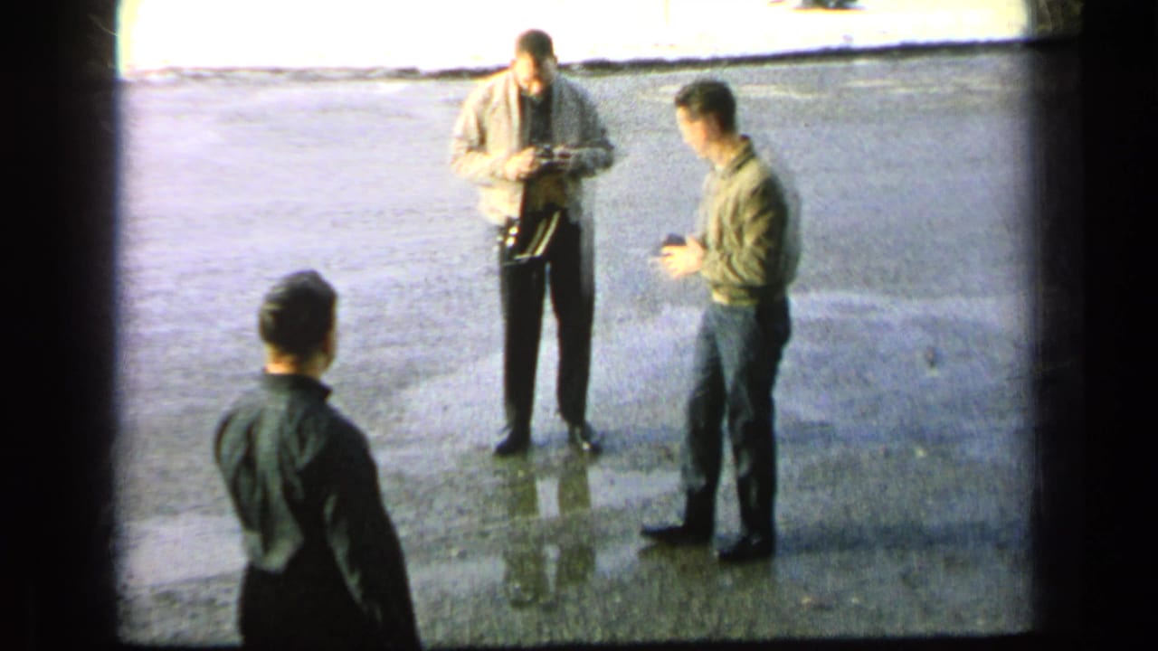 1963 Germany military-base home-movie frame showing adult men on reflective pavement, with one man in a light jacket handling a camera.