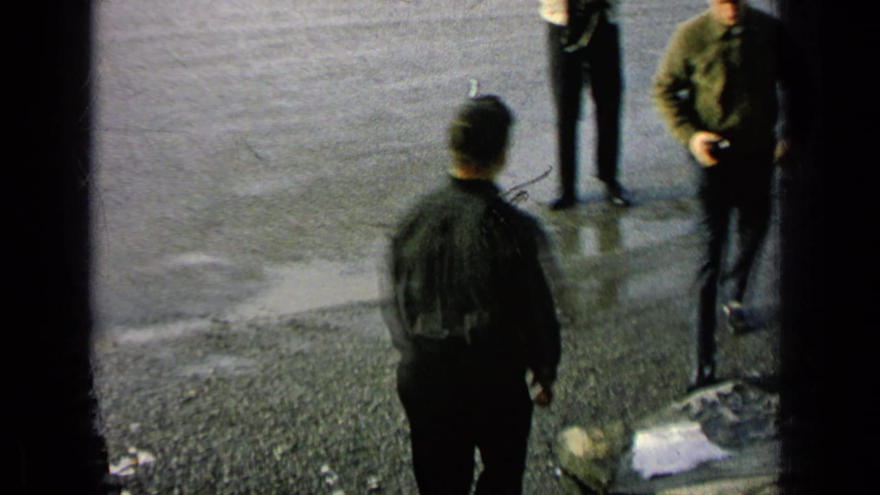 Adult men stand on wet pavement, with one central figure seen from behind and others partly cropped.