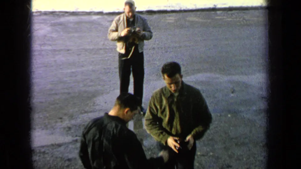 1963 8mm frame showing men on a wet Germany military-base surface, including a man in a light jacket adjusting a camera.
