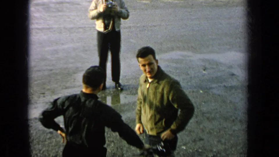 Home-movie frame with adult men at a Germany military-base setting, one looking toward the camera as others handle photographic gear.