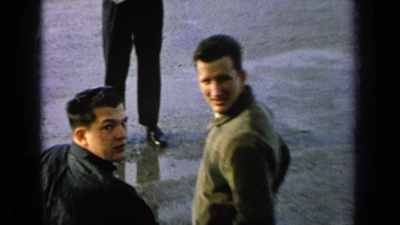1963 8mm frame from a Germany military-base reel showing young men in casual clothing looking up in a paved outdoor area.