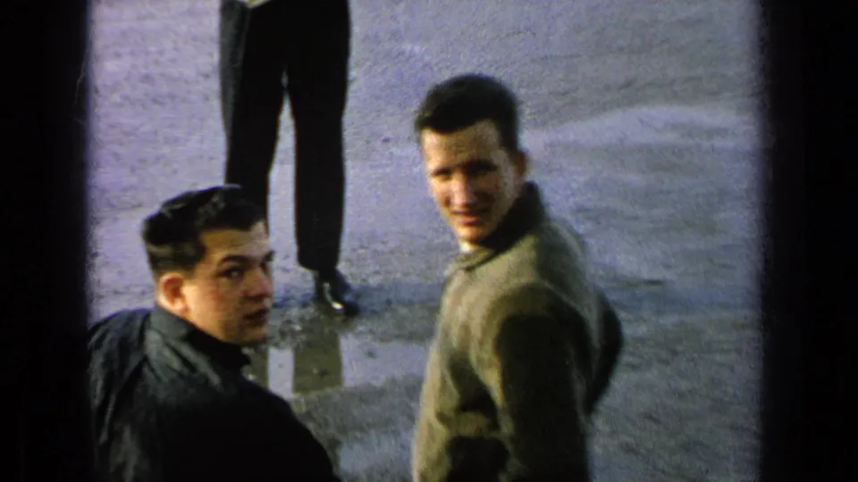 1963 8mm frame from a Germany military-base reel showing young men in casual clothing looking up in a paved outdoor area.