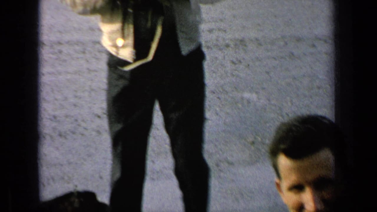 A partial figure with a camera strap stands near the edge of a paved lot as another man is partly visible.