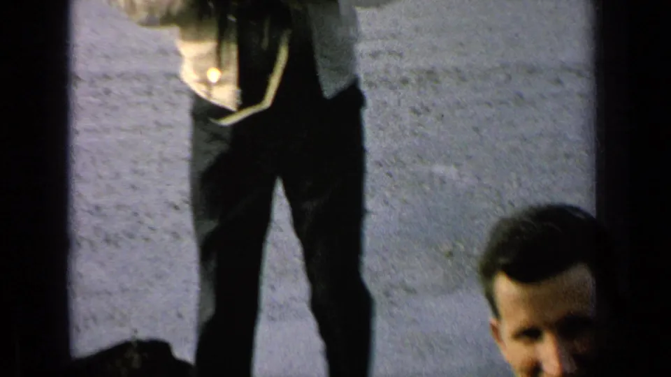 A cropped 1963 8mm view shows a man holding or wearing a camera near a paved lot, with another face partly entering the frame.