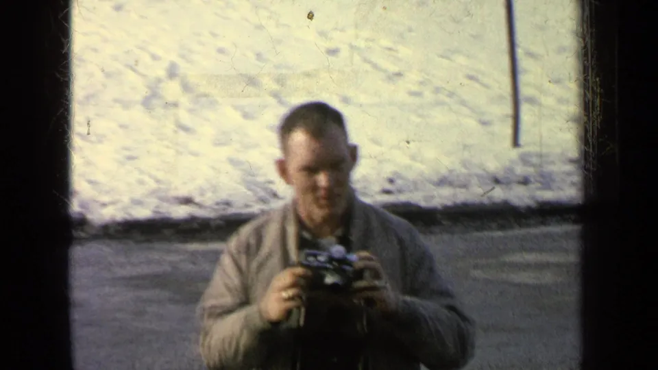 1963 8mm frame from the Germany military-base reel showing a man in a light jacket holding a camera near snow and pavement.