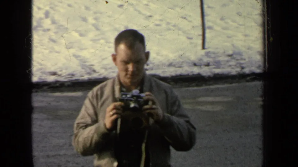 The frame shows a man in a light jacket handling a camera beside a snow-covered curb in 1963 Germany military-base home-movie footage.