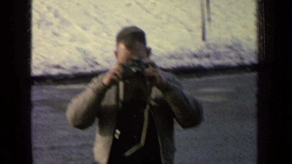 A man in a casual jacket films near a snow-covered road in 1963 Germany military-base home-movie footage.