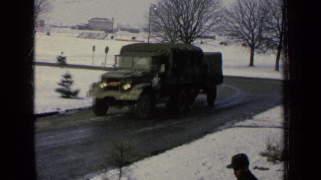 Green military cargo truck driving on a curved snowy road with bare trees nearby.