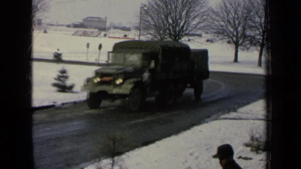 A green U.S. Army-style cargo truck drives along a snow-lined road on a German base in 1963 footage.