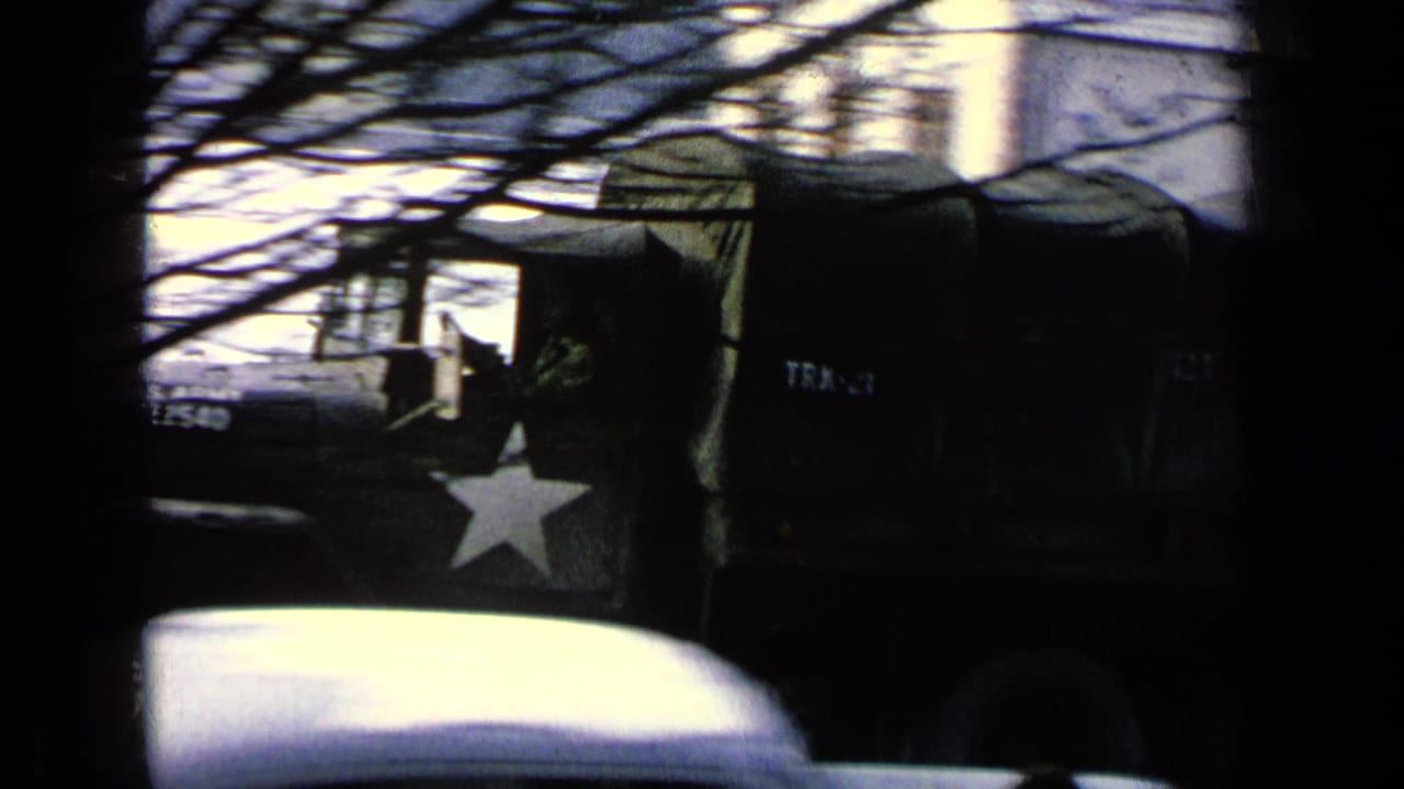 1963 8mm frame of a dark military truck on a base road, partly hidden by tree branches with a white star insignia visible.