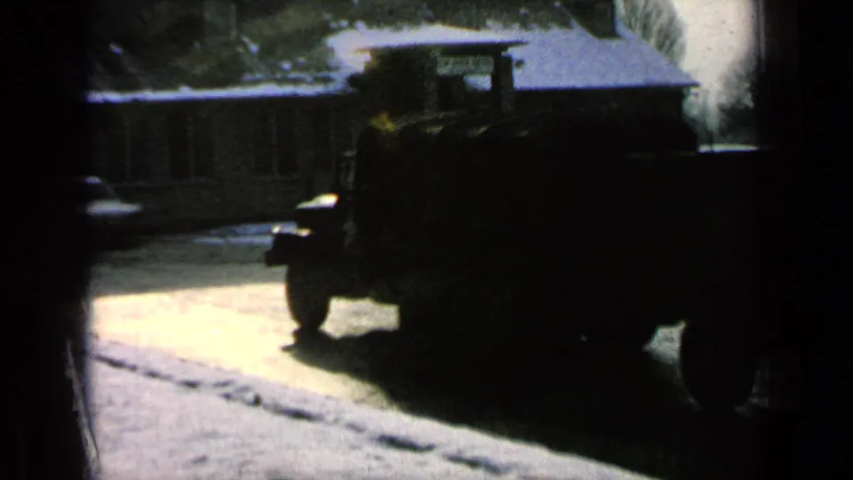 Dark silhouette of the military truck passing along a base road, with winter trees and a low building visible behind it.