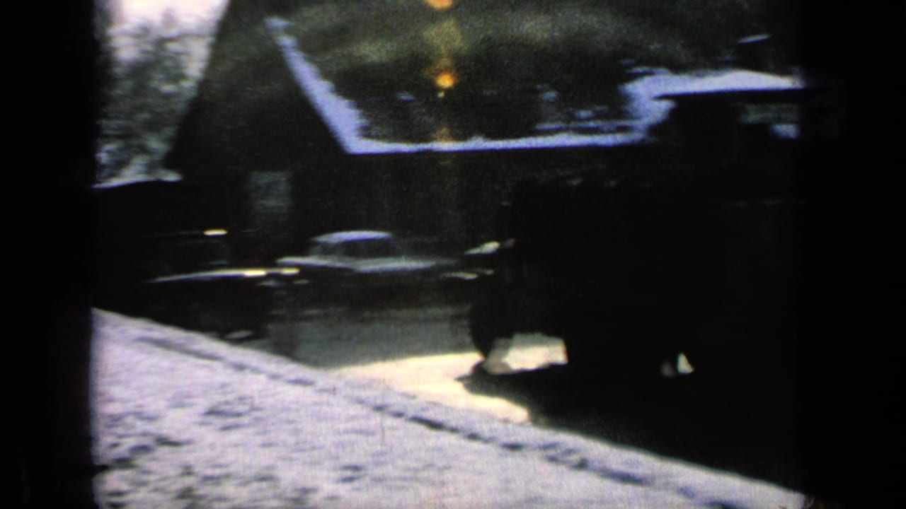 Snowy street with houses, bare trees, and dark vehicles seen through heavy film grain.