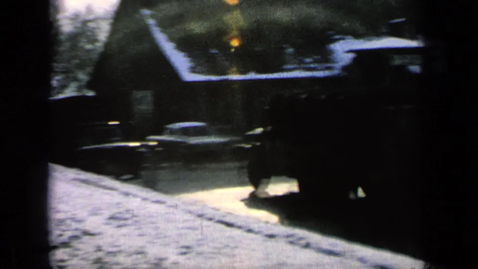 1963 home-movie frame of a snowy street near a German military base, with houses, bare trees, and vehicles partly obscured by exposure and grain.
