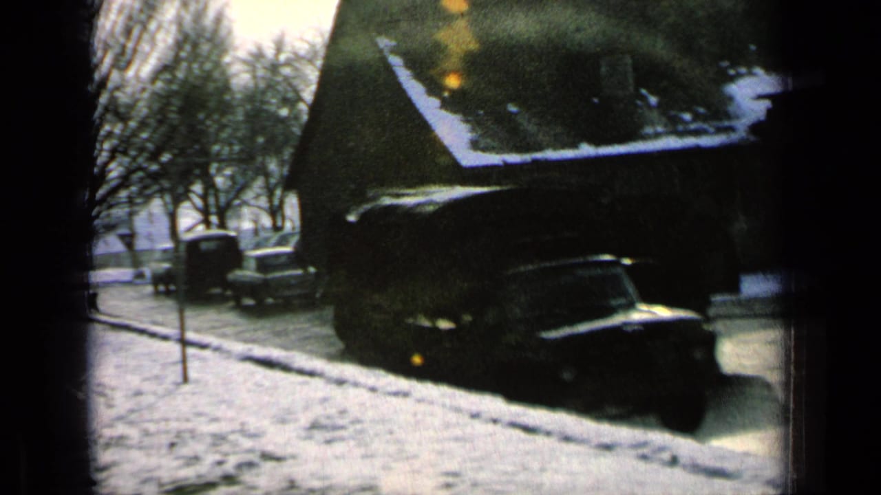 Snowy roadside with a parked car, bare trees, houses, and a vertical street sign.