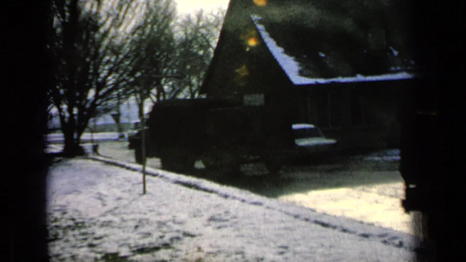 1963 home-movie view of a snow-covered street in Germany, showing a dark vehicle near houses, bare trees, and a roadside sign.