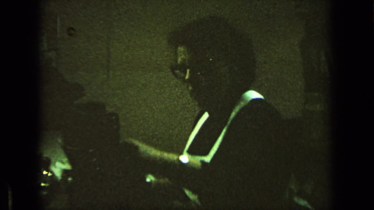 Green-tinted 8mm frame shows a man in glasses leaning over a desk or writing surface indoors.