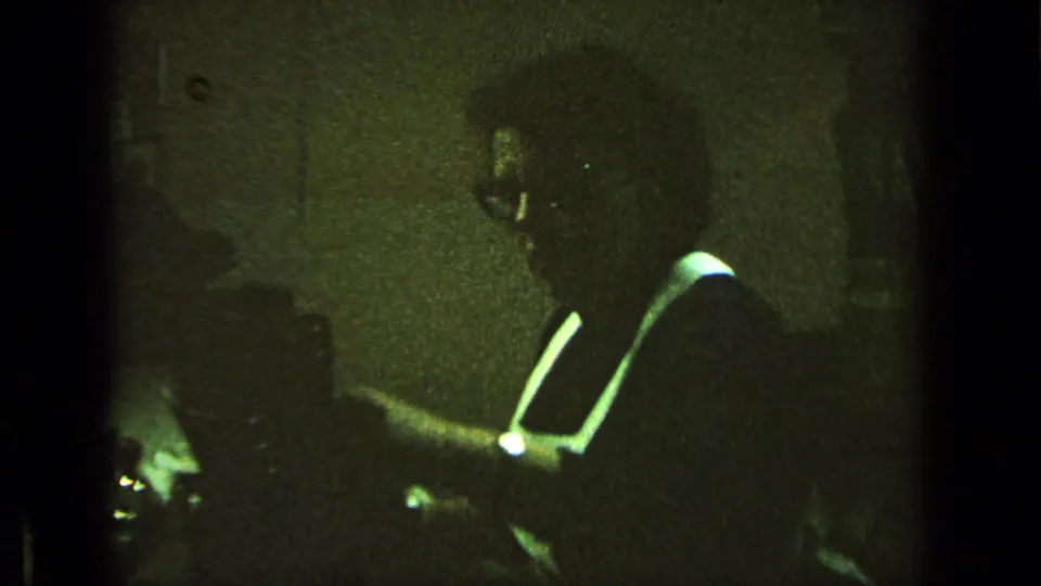 Green-tinted 8mm frame shows a man in glasses leaning over a desk or writing surface indoors.