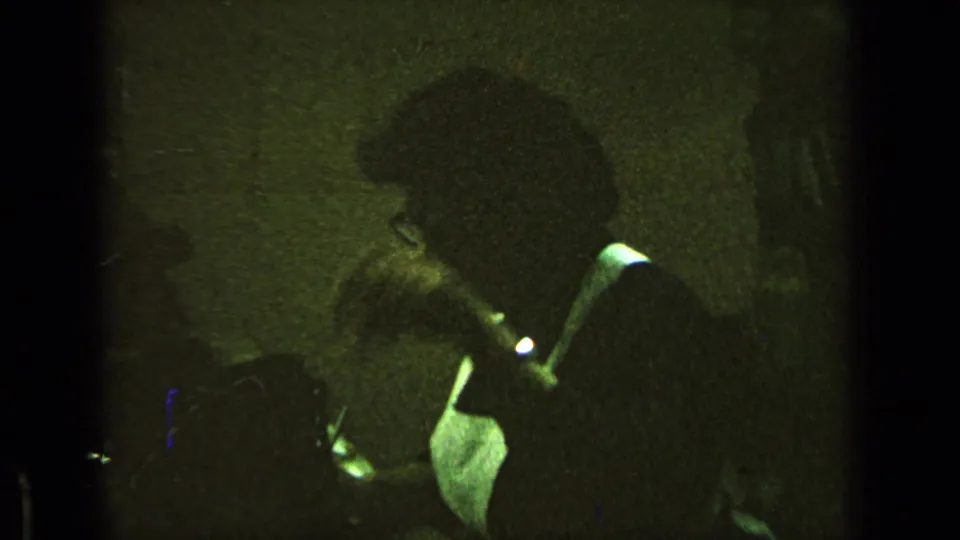 A dim 8mm interior view shows a man with glasses leaning toward papers or a desktop.