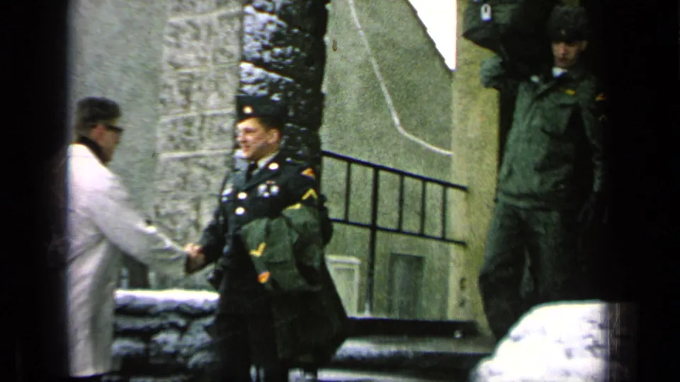 A civilian in a light coat shakes hands with a uniformed soldier outside a stone base entrance as another soldier stands nearby.