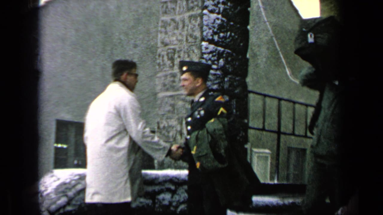 At a 1963 military base in Germany per metadata, a uniformed soldier shakes hands with a man in a white coat near snowy stone steps.
