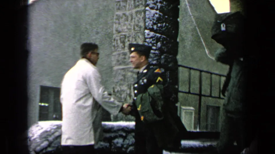 At a 1963 military base in Germany per metadata, a uniformed soldier shakes hands with a man in a white coat near snowy stone steps.