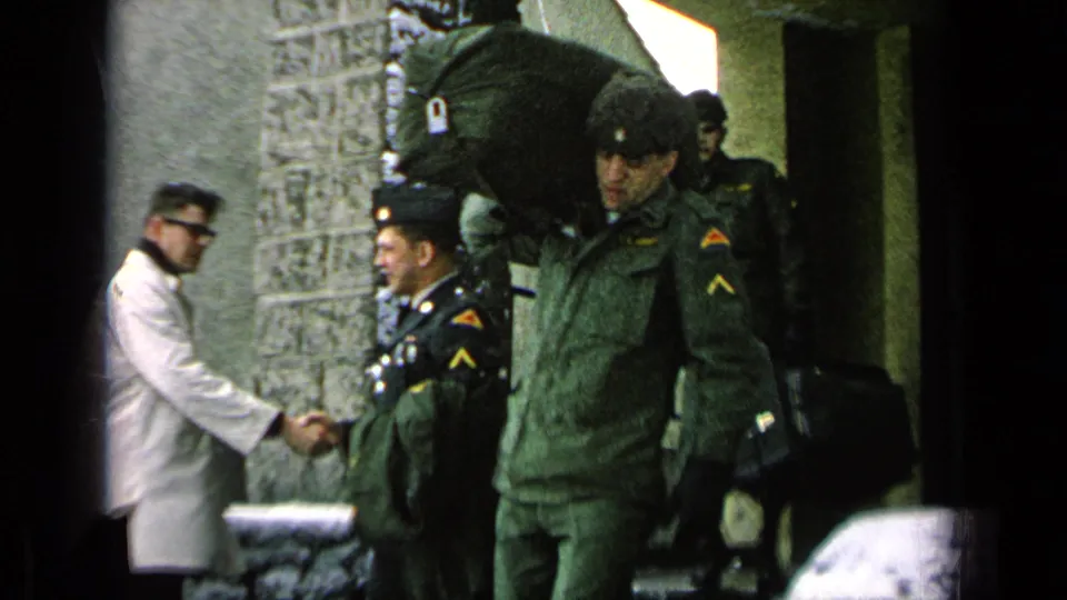 A soldier carrying a large duffel bag stands by as another soldier shakes hands outside a snowy base building in Germany, per metadata.