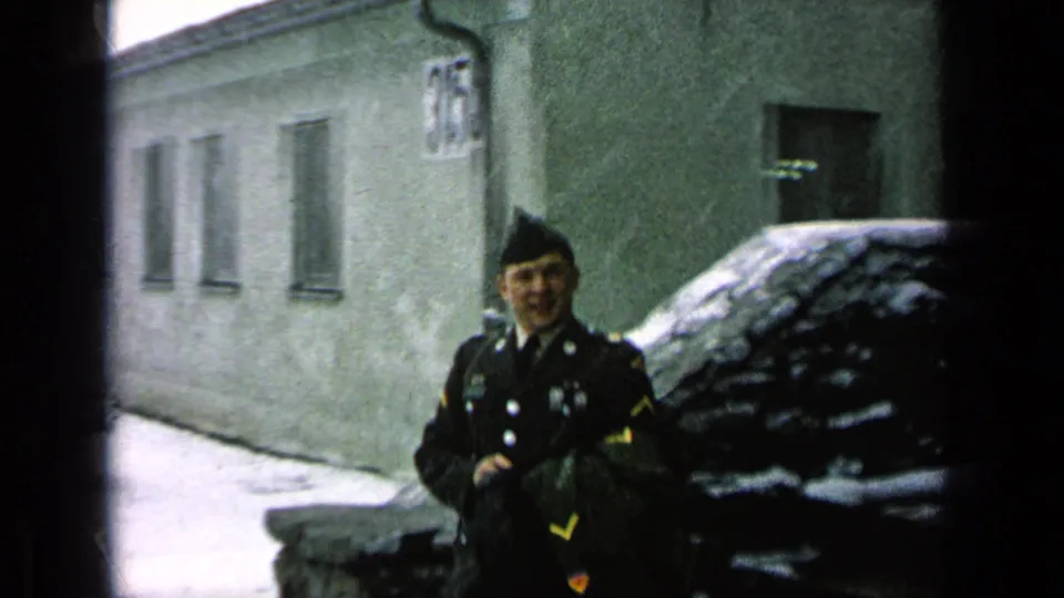 A uniformed soldier smiles toward the camera in a snowy base area, with a barracks-like building and partially snow-covered car behind him.