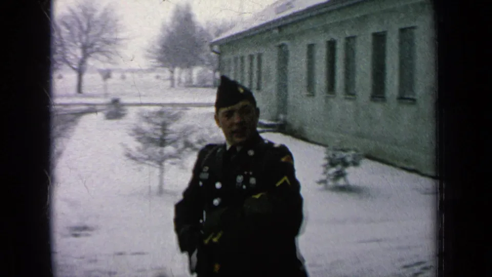 A uniformed soldier stands on a snowy walkway at a military base in Germany, with a long barracks-like building and bare winter trees visible.