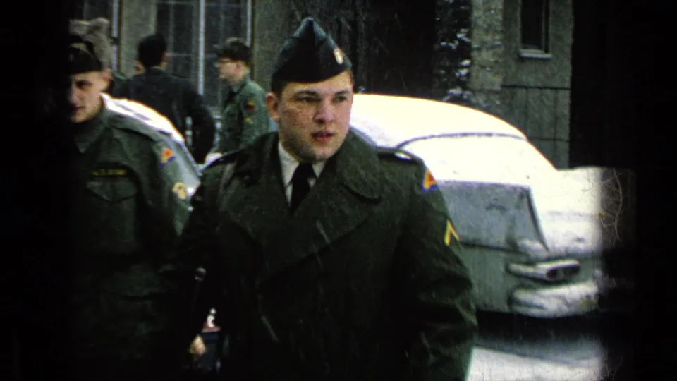 Young U.S. Army soldiers in olive drab uniforms stand near a snow-covered car in archival home-movie footage from a German base setting.