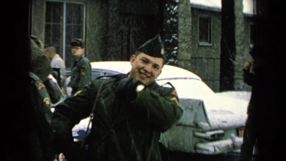 A uniformed soldier smiles and waves toward the camera while standing near other servicemen and a snow-covered car.