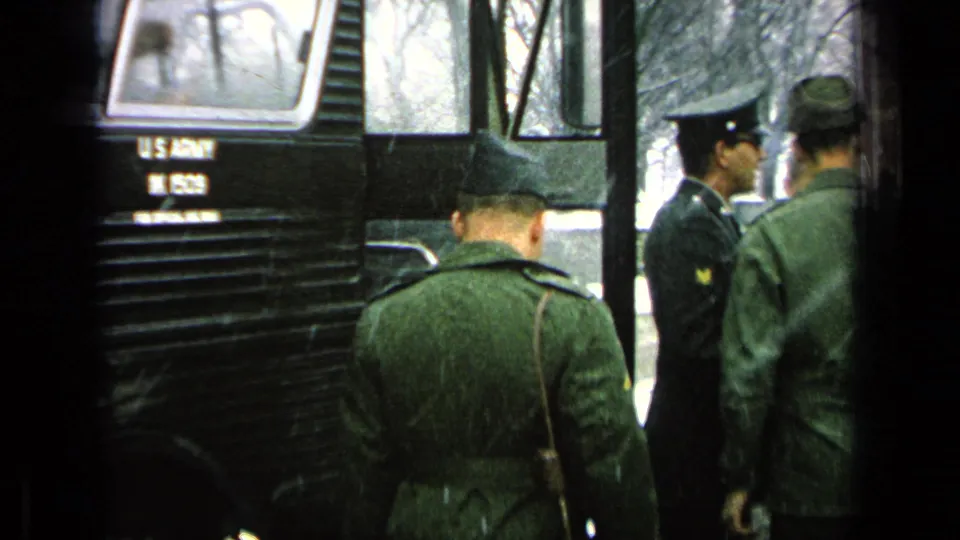 Uniformed soldiers gather beside a dark military bus marked for U.S. Army use, with snow and bare trees visible in the background.