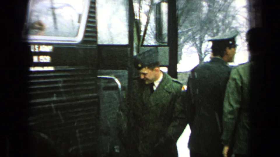 Uniformed soldiers pass the side of a dark U.S. Army bus marked M 1508, with faces visible.