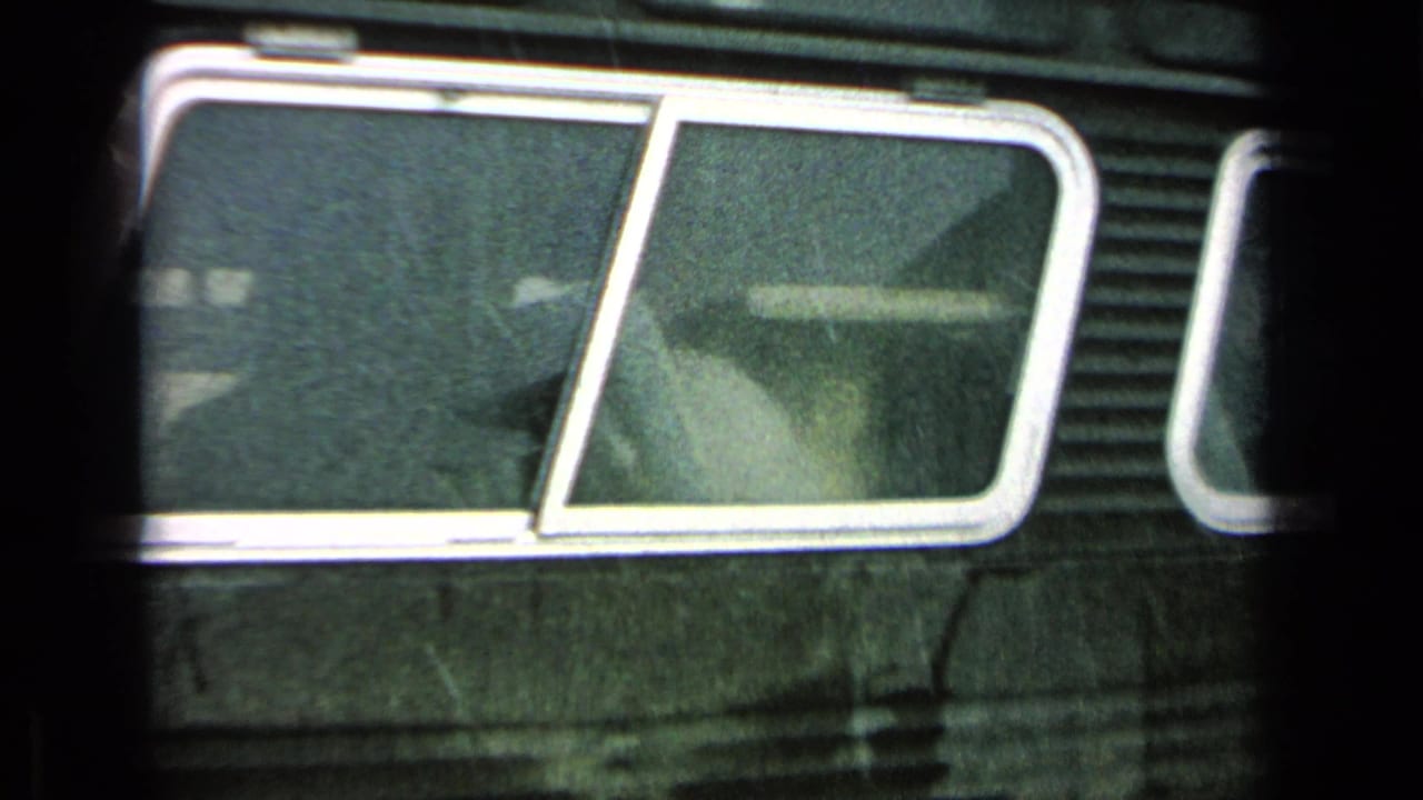 Close 8mm view of a dark Army bus window area, showing rain, reflections, and a faintly visible passenger or interior shape.