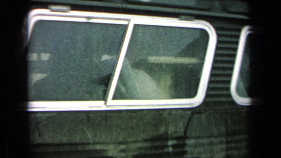 Close 8mm view of a dark Army bus window area, showing rain, reflections, and a faintly visible passenger or interior shape.