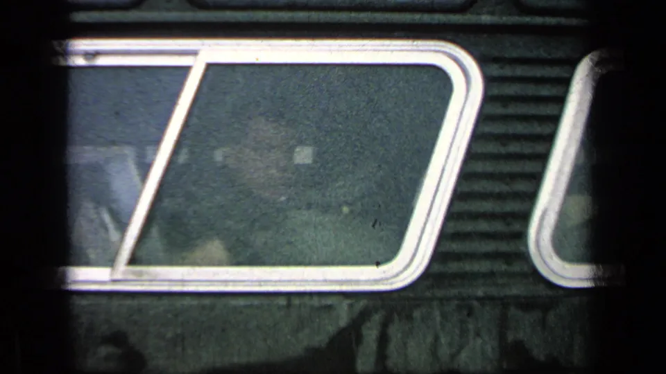 Rainy close-up of a bus window on dark ribbed siding, with soft reflections and little detail visible through the glass.