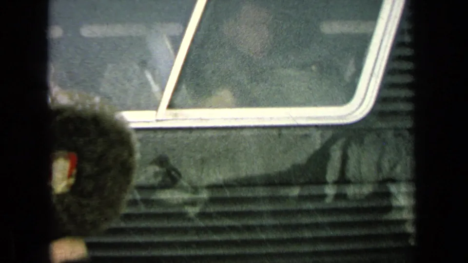 Home-movie frame of the Army bus side in rain, with a blurred dark-uniformed person partly visible below the window.